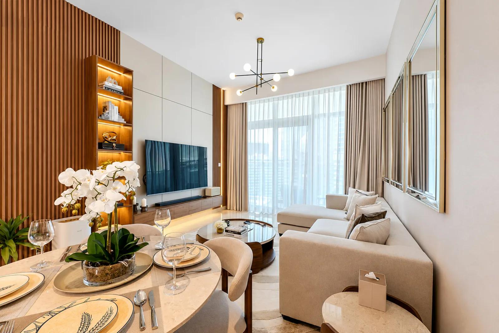 Bright apartment living and dining room with beige seating, a wood slat wall, television, orchids, and large windows.